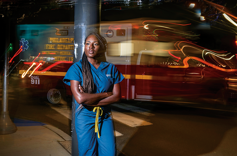 Chidinma Nwakanma, MD, wearing dark blue scrubs, stands outside at night with her arms crossed while a motion-blurred ambulance passes behind her.